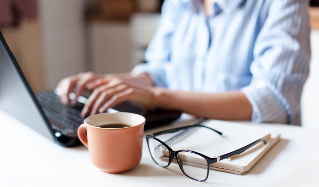 Person sitting at desk typing on a laptop. Coffee, glasses and notepad next to laptop.