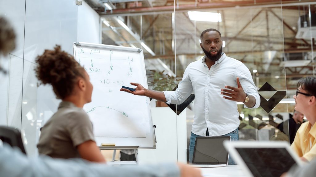 Young afro american businessman pointing at white blackboard and explaining new project to his colleagues while working together in the creative office. Teamwork. Presentation