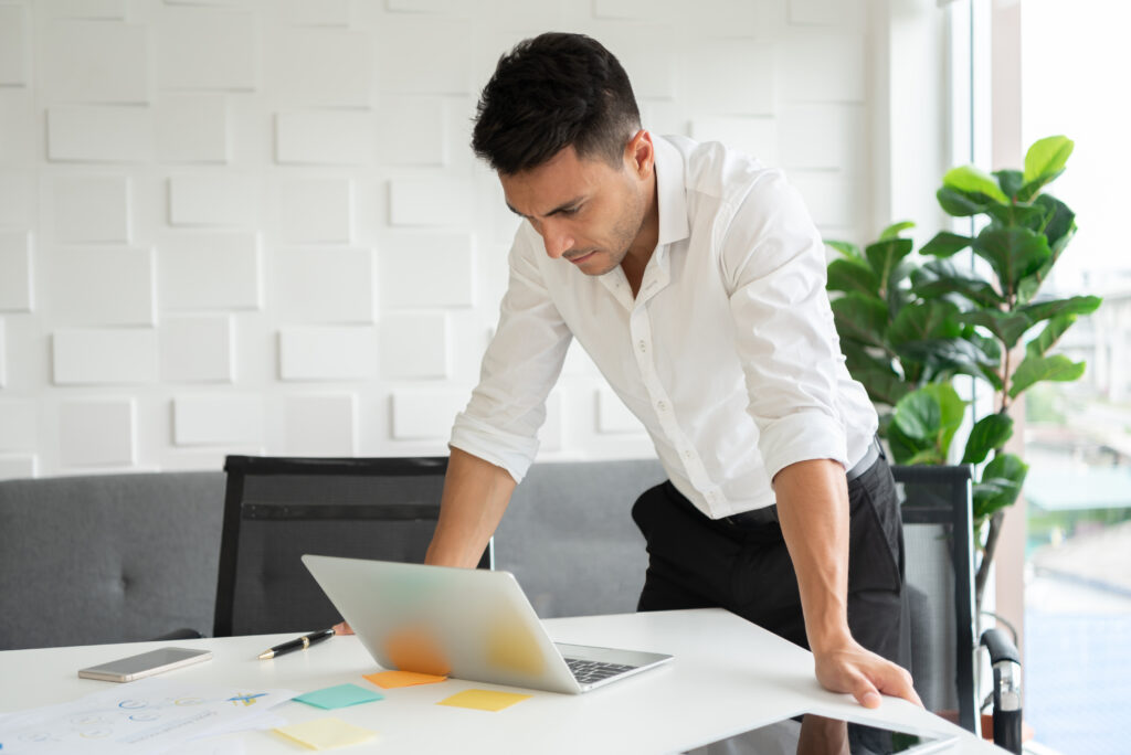 Young businessman is standing with hands on desk, looking at a laptop.