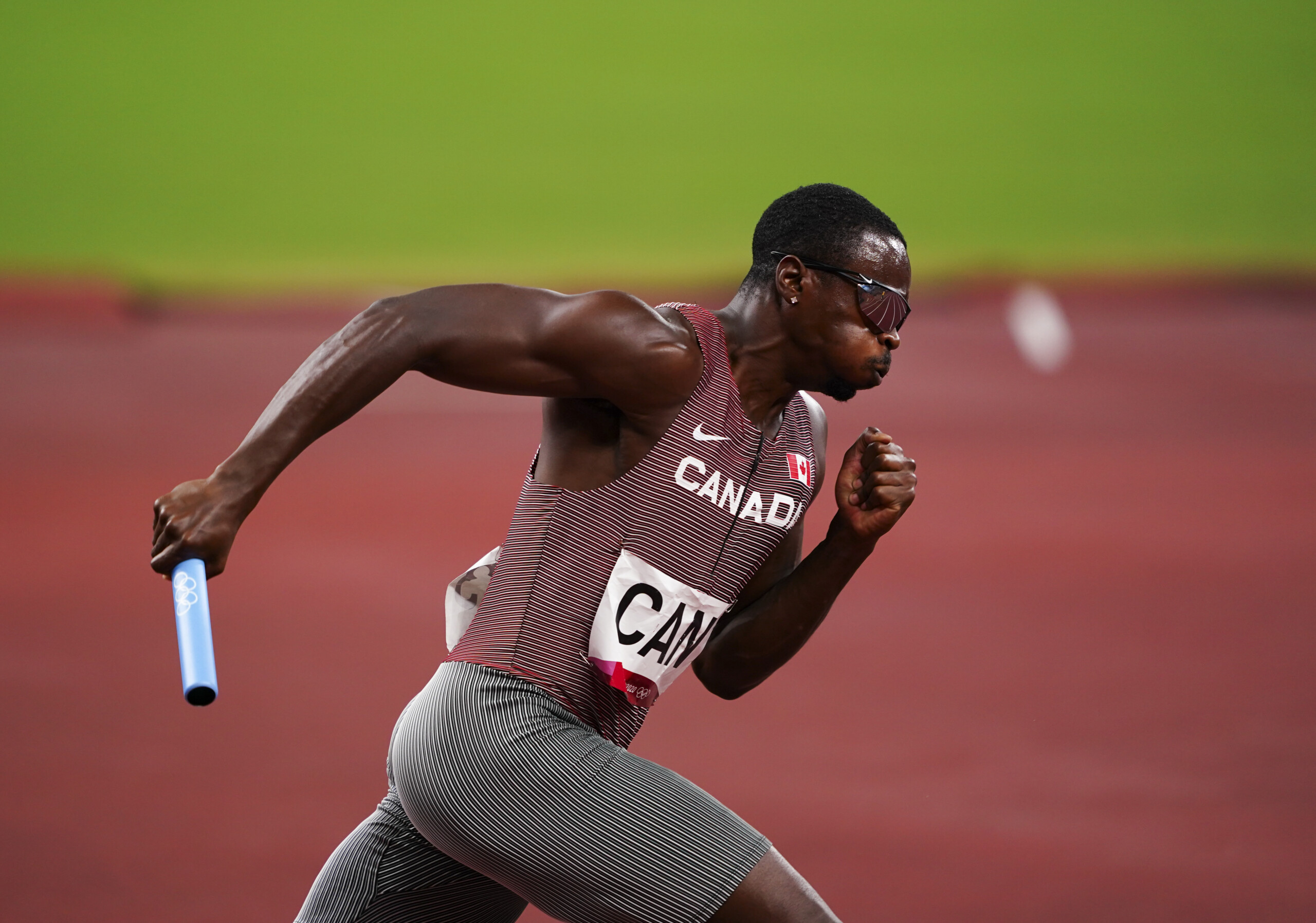 A black athlete wearing a red and white tank top with grey short tights is sprinting in a race with a baton in his right hand.